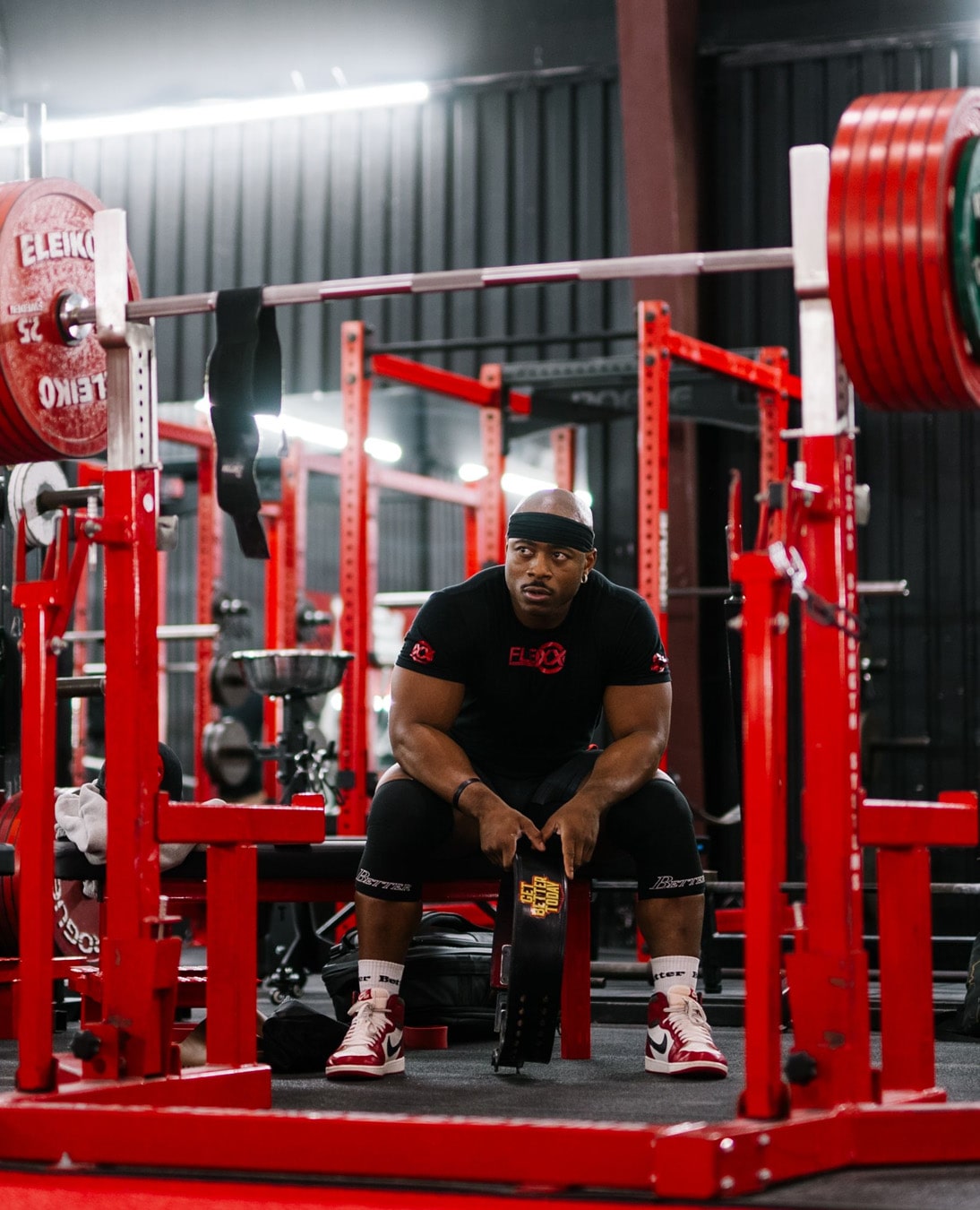 A man sits on a bench in a gym with weightlifting equipment around him.