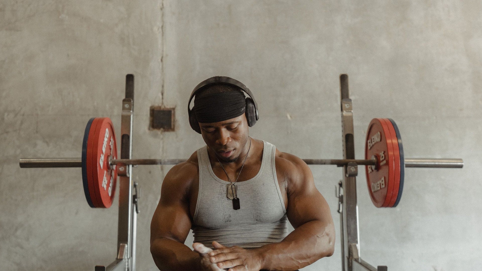 Man in gym preparing to lift weights, wearing headphones and a gray tank top.
