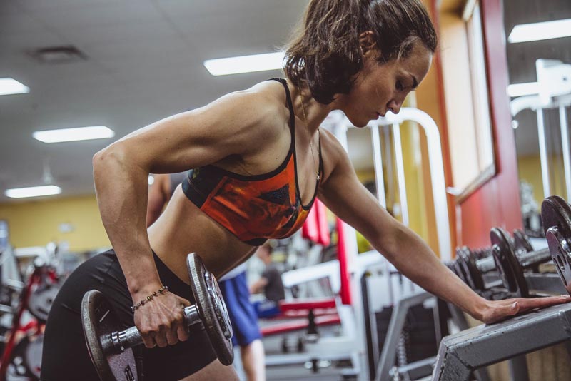 A woman with brown hair pulled in a ponytail, wearing a red sports bra and black shorts is lifting a dumbbell.